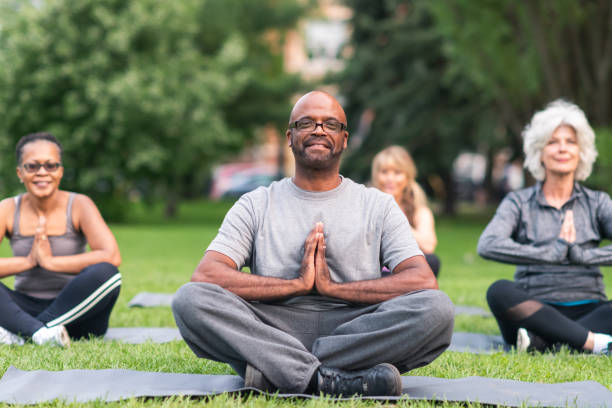 A multi-ethnic group of seniors is attending a yoga class outdoors. The group is sitting on yoga mats. They are meditating. The individual in focus is a black man. He is sitting at the front of the group. He is smiling directly at the camera.